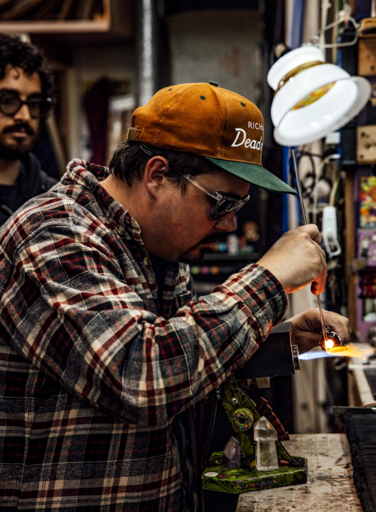 Young man blowing glass in workshop with onlooker in background